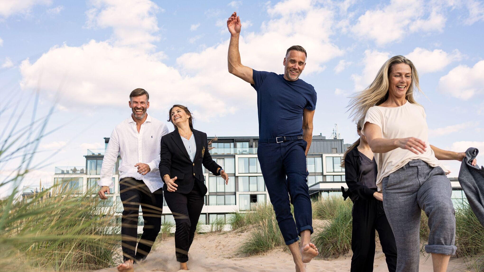Fem glada personer springer i sanddyna med Hotel Tylösand i bakgrunden