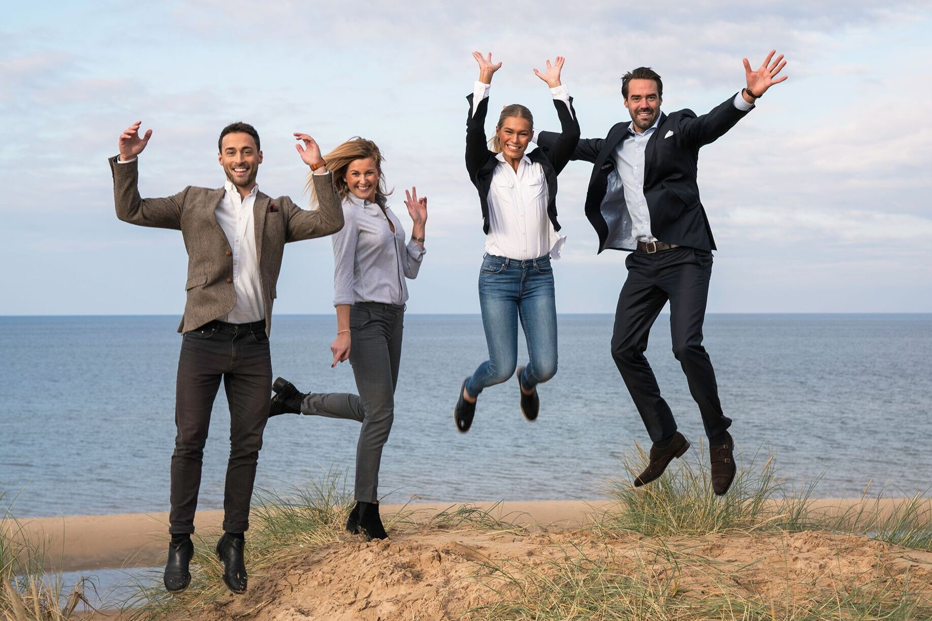 Fyra glada personer hoppar i sanddyna med havet i bakgrunden